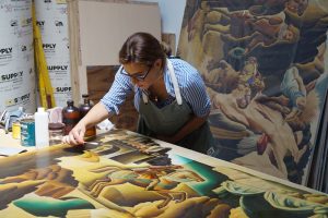 a female art conservator applies a treatment to a mural that is laid out on a table