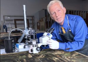 an older, well-dressed white man with gloves on using a microscope to analyze a painting