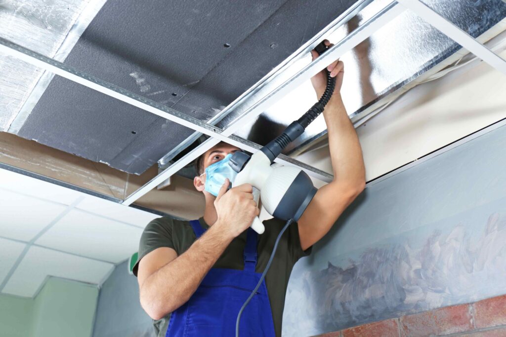 a man wearing a mask uses an industrial vacuum to clear dust from a hard-to-reach surface